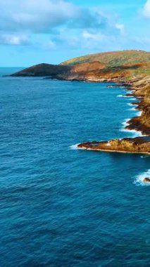 Karanlık volkanik kayaların bulutlu bir gökyüzünün altında parlak mavi okyanus dalgalarıyla buluştuğu sivri bir kıyı şeridi. Hawaii 'de engebeli kayalık kıyı ve mavi okyanus suyu. Dikey video.