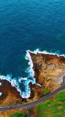 Beyaz sörfün havadan görünüşü bir sahil otoyolunun yanındaki koyu kahverengi kayalık bir noktaya çarpıyor. Dalgalar volkanik kayalara çarpıyor. Dikey video.