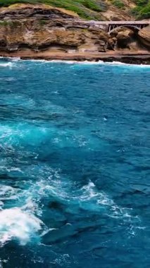 Katmanlı kaya uçurumunun önünde beyaz köpüklü mavi okyanus suyu ve taş kemer köprüsünün havadan görünüşü. Okyanus dalgaları bir kaya duvarına çarpıyor. Dikey video.
