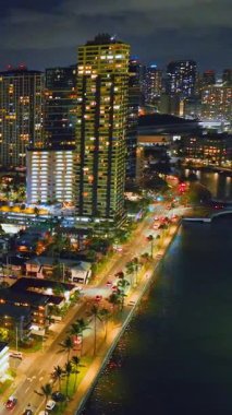 Geceleri kıyı yolu ve suyun yanında aydınlık gökdelen olan bir gökdelenin havadan bakış açısı. Honolulu, Waikiki 'de yüksek bir binanın gece görüntüsü. Dikey video.