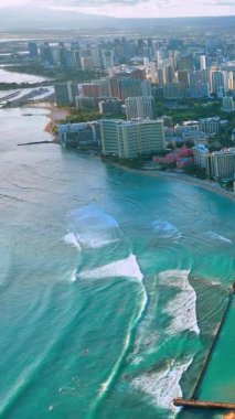 Okyanus dalgaları kıyıya yaklaşıyor. Şehir binaları ve arka planda gün ışığında kıyı şeridi var. Waikiki sahilindeki dalgaların yüksek açılı hava görüntüsü. Dikey video.