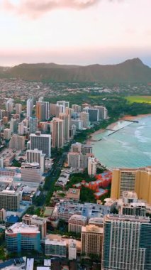Şehir binalarının yüksek açılı manzarası ve yumuşak bir akşam gökyüzünün altında dağ silueti olan kıyı şeridi. Honolulu ve Diamond Head üzerinde günbatımı görüntüsü. Dikey video.