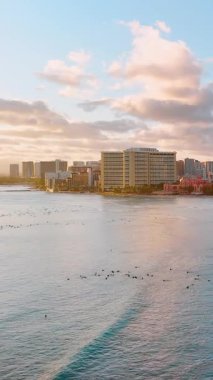 Okyanusta sörfçüler Honolulu silueti ve bulutlu bir gökyüzünün altında pembe otel mimarisi. Hava görüntüsü. Dikey video.