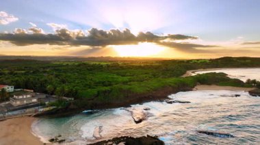 Gökyüzünü ve okyanus dalgalarını aydınlatan yeşil bitki örtüsü ve güneş ışınlarıyla kayalık bir kıyı şeridinde gün batımı. Güneş ışınları tropikal kıyılarda bulutları delip geçiyor..