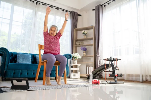Senior woman sitting on chair and raising two arms above her head. doing workout for good healthy with training exercise online on tablet, elderly workout online from home concept