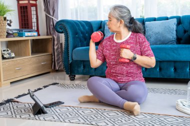 Portrait retirement woman sitting on mat in the living room, She holding dumbbell and  lifting on shoulder, Senior workout during quarantine concept