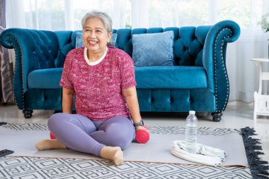 Portrait retirement woman sitting on mat in the living room, She smile and Holding dumbbell in hand, Senior workout during quarantine concept