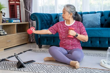Portrait retirement woman sitting on mat in the living room, She holding dumbbell and lifting extend arms to sides, Senior workout during quarantine concept