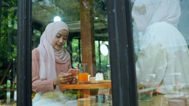 Cam pencerenin yanında oturan genç ve güzel Müslüman kadın ve arkadaşının olduğu cam camdan bakıp akıllı telefon kullanıyorlar ve kafede sohbet ediyorlar.