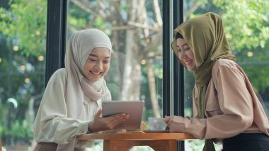 Portreli neşeli Müslüman kadın ve tesettürlü arkadaşı tabletle internetten alışveriş yapıyorlar, kafeteryada mutlulukla birlikte gülüyorlar, müslüman işi konsepti.