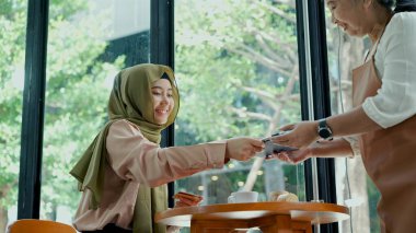 Seçici odak, tesettürlü genç, tesettürlü güzel müslüman kadın kahve dükkanında cam pencerenin yanında oturuyor kredi kartıyla yaşlı garsona ödeme yapıyor. 