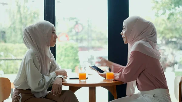 年轻美丽的穆斯林妇女和戴头巾的朋友坐在玻璃窗边 在咖啡店里用智能手机聊天 这是穆斯林商人的工作理念 — 图库照片