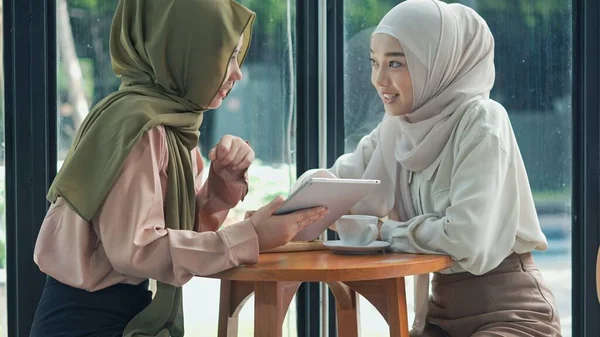 Portrait cheerful muslim woman and her friend wearing hijab are shopping online with tablet,  They are smiling together with happiness in coffee shop, muslim business working concept