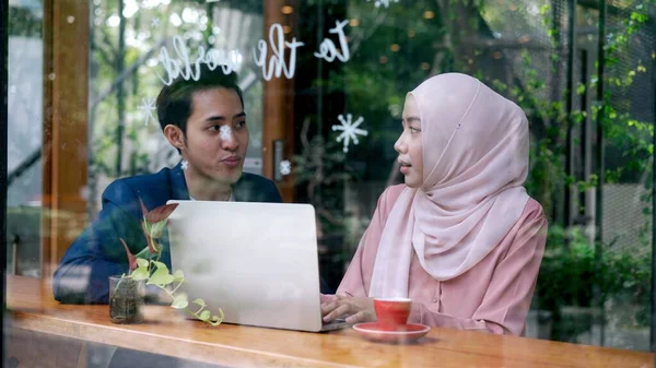 窓ガラスを通して見る ラップトップ上で働くヒジャーブを身に着けているイスラム教徒の男性と若い女性 彼らは一緒にコーヒーショップで議論しています イスラム教徒のビジネスワーキングコンセプト — ストック写真