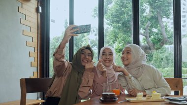 Kafede cam pencerenin yanında oturan tesettürlü bir grup güzel Müslüman kadın mutlulukla selfie çekmek için akıllı telefon kullanıyorlar.