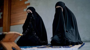Genç Müslüman kadın ve kız kardeş geleneksel siyah giysiler giyer, paspasa oturur, Tanrı 'ya dua eder, evde el ele dua eder.