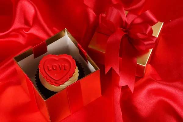 Cupcake with a red heart on top in gifts boxes on red satin back — Stock Photo, Image