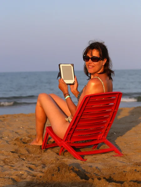 Kobieta na czerwone krzesło odczytuje ebook na plaży nad brzegiem morza w su — Zdjęcie stockowe