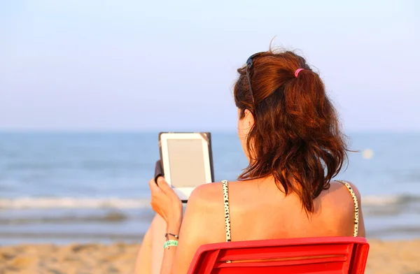 Ładna kobieta na czerwone krzesło odczytuje ebook na plaży latem — Zdjęcie stockowe