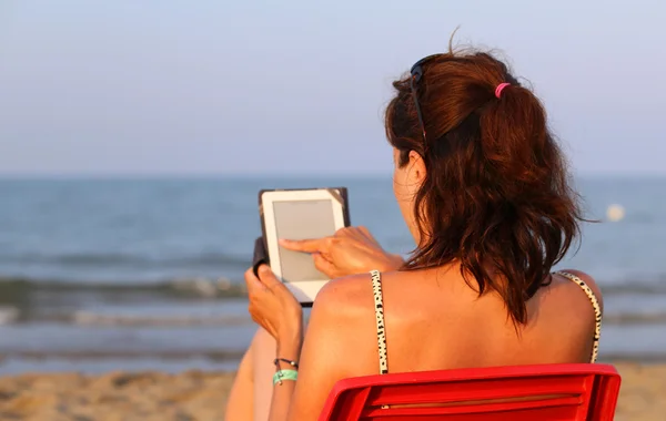 Donna sulla sedia rossa legge l'ebook sulla spiaggia in riva al mare — Foto Stock