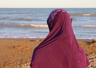 Yazın gün batımında deniz kenarında başında mor peçeli bir Arap kadın.