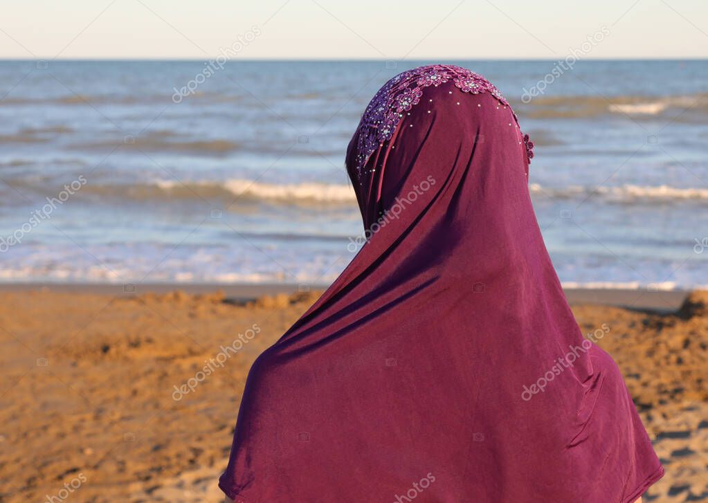 Arab woman with a purple veil on her head by the sea at sunset in summer
