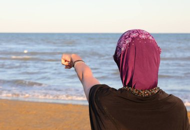 Saçını örtmek için duvağı olan genç bir kadın deniz kıyısında işaret parmağıyla ufku gösteriyordu.