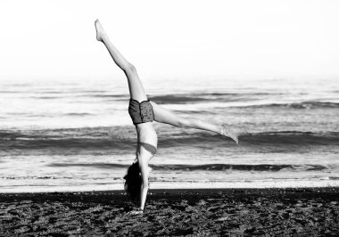 Genç atletik kız, sahilin kenarında siyah beyaz efektlerle tekerlek yaparak jimnastik yapıyor.