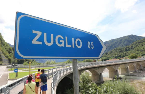 川の上の橋を通ってイタリアのZuglioと呼ばれる小さな村に到達する方向と道路標識 — ストック写真
