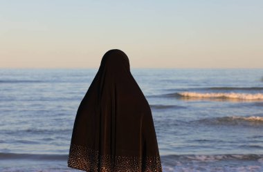 Genç Arap kadın, sakin denizin kenarında siyah elbiseli, saçlarını örten.