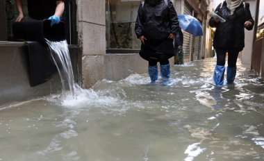 İşçi dükkanındaki suyu kovayla boşaltıyor ve diğer insanlar sokakta yürürken su geçirmez tozluklarıyla Venedik İtalya 'daki yüksek gelgit yüzünden sular altında kalıyor.