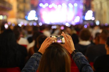 Açık hava konserinde akıllı telefonuyla video çeken bir kız. Bir sürü seyirci ve sahne ışıl ışıl.