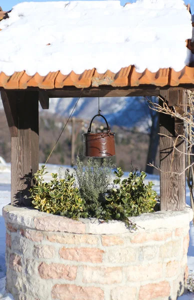 Pozo con cubo y nieve en el techo en pueblo de montaña —  Fotos de Stock