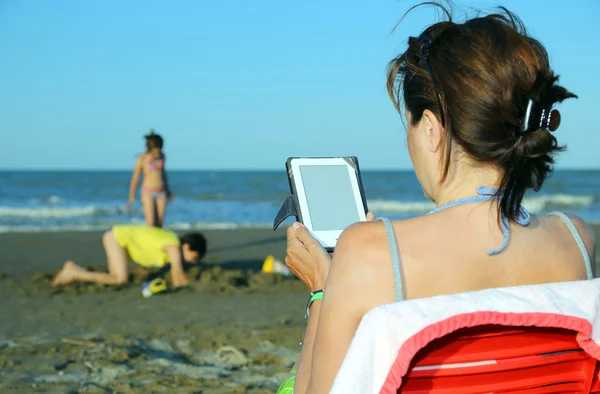 Donna legge l'ebook sulla spiaggia mentre i suoi figli giocano — Foto Stock