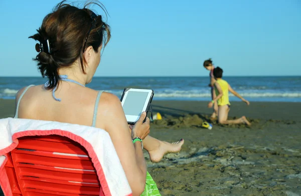 Kobieta czyta ebook podczas jej dzieci bawiące się na plaży — Zdjęcie stockowe