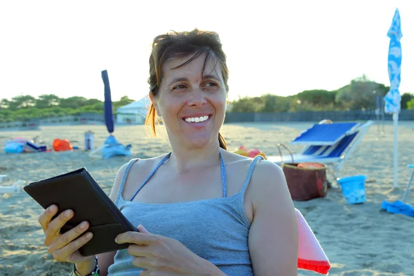 Vackra leende kvinna läser ebook på stranden — Stockfoto