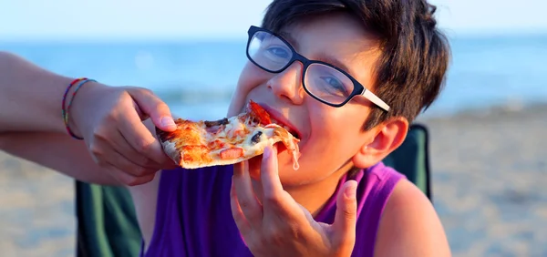 Młody chłopak zjada kawałek pizzy na plaży o zachodzie słońca latem — Zdjęcie stockowe