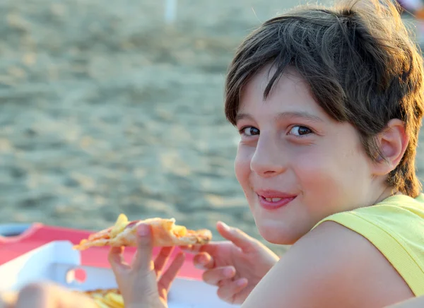 Un garçon souriant mange une pizza avec des chips sur la plage — Photo