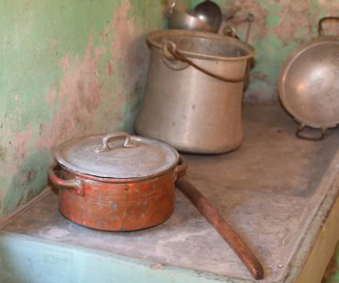 Antika metal pişirme tencereleri ve tavaları aşınmış yeşil duvar arka planına karşı sade ahşap bir rafa konur.