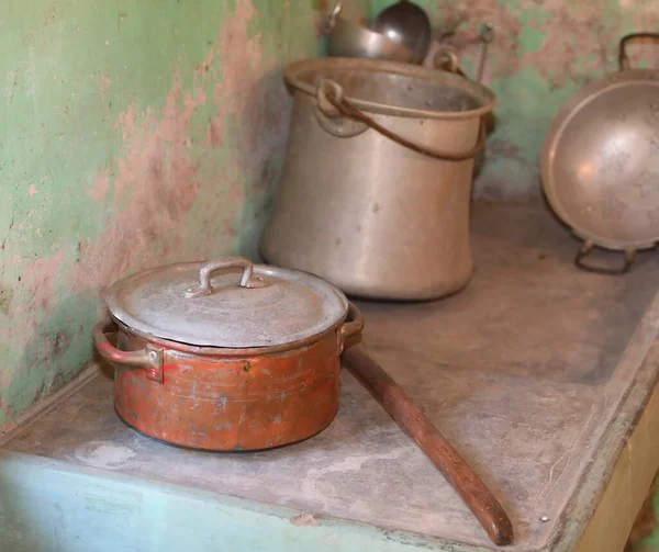 Antika metal pişirme tencereleri ve tavaları aşınmış yeşil duvar arka planına karşı sade ahşap bir rafa konur.