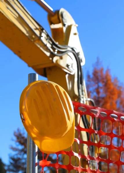 Chapéu duro amarelo no canteiro de obras da estrada e uma escavadeira — Fotografia de Stock
