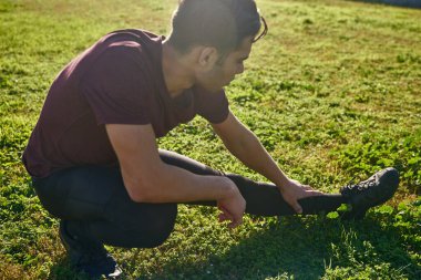 Çimlere çömelmiş genç adam kaslarını esnerken ön bacağını tutuyor.
