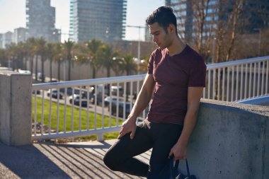 Sıcak güneş ışığında bir köprüde duran adam spor çantasını tutarken aşağıya bakıyor.