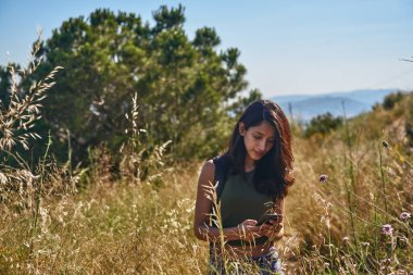 Vahşi bitki örtüsüyle çevrili vahşi doğada dikilirken dikkati dağılmış bir kadın mesaj atıyor.