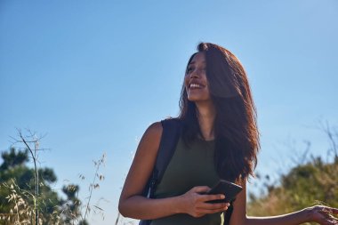 Açık havada neşeli bir bayan bir eliyle telefonunu tutarken diğer eliyle vahşi florayı nazikçe okşuyor.