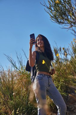 Açık güneşli bir günde, vahşi bir bitki örtüsüyle çevrili bir tepede selfie çeken havacı genç bayan.