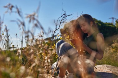 Etrafı vahşi bitki örtüsüyle çevrili bir kayanın üzerinde oturan dikkati dağılmış genç bayan.