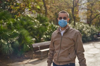 Ağaçlarla çevrili bir parkta dikilip doğrudan kameraya bakan orta yaşlı bir adam.