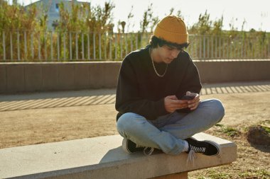 Gün boyunca bankta tek başına otururken cep telefonu kullanan çapraz bacaklı beyaz bir adam.