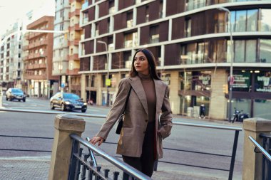 Merdivenlerin tepesinde duran başka bir kadın nazikçe korkuluklara tutunuyor ve elini cebine sokuyor.